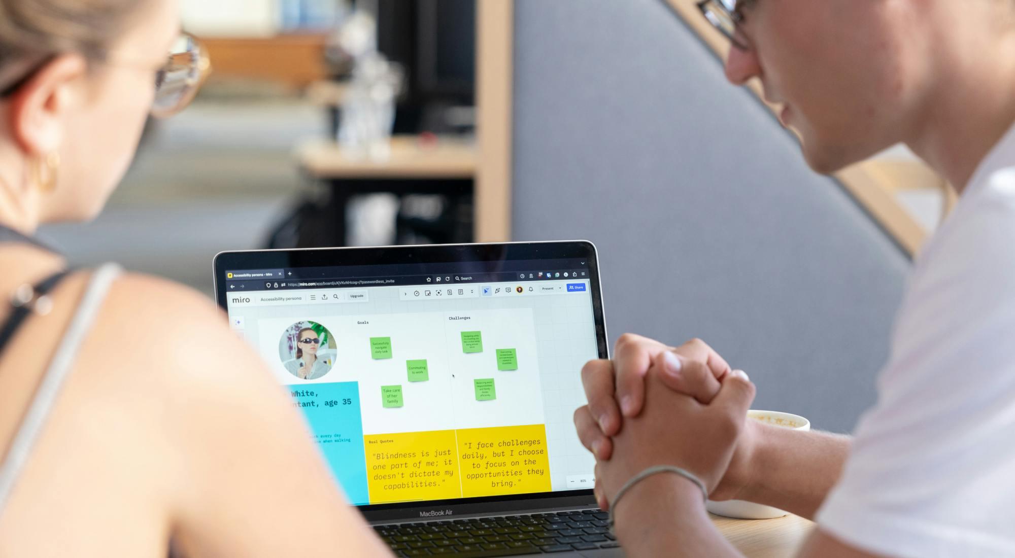 Two individuals sitting side by side are focused on a laptop screen displaying a Miro board with various notes and a persona profile. One person is wearing glasses and both are engaged in a collaborative discussion.